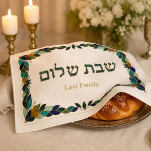May include: A rectangular off-white fabric challah cover featuring a decorative border of blue, green, and gold leaf-like patterns. The Hebrew text "שבת שלום" is printed in dark green, with the English text "Levi Family" printed below in gold.