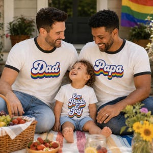 May include: Two white ringer t-shirts with black trim, one featuring the blue text Dad and the other Papa, both with a rainbow-coloured wavy underline. A matching white t-shirt for a child features the blue text Our Pride & Joy with a rainbow design.