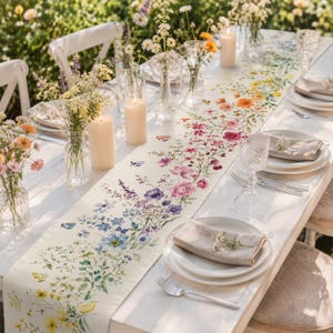 May include: A long rectangular table runner featuring a colorful botanical print of wildflowers, greenery, and butterflies on a cream background, displayed on a white wooden table with place settings, glass vases, and pillar candles.