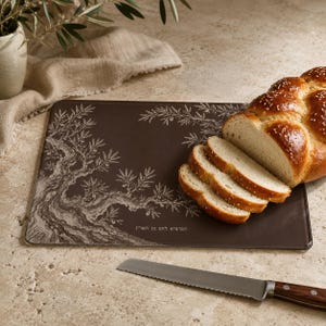 May include: A rectangular dark brown glass cutting board featuring a white etched olive tree design and Hebrew text. A loaf of braided challah bread is sliced on the board, accompanied by a serrated bread knife with a wooden handle.