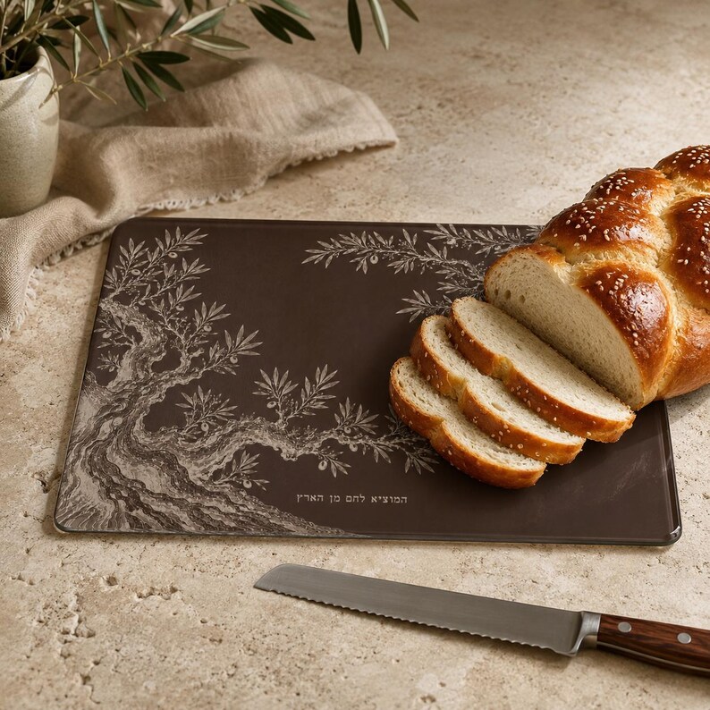 May include: A rectangular dark brown glass cutting board featuring a white etched olive tree design and Hebrew text. A loaf of braided challah bread is sliced on the board, accompanied by a serrated bread knife with a wooden handle.