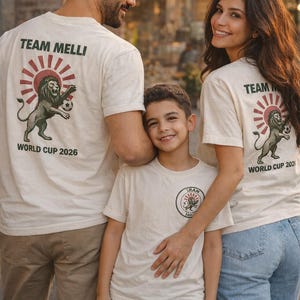 May include: Off-white short-sleeve t-shirts featuring a graphic on the back with a lion holding a soccer ball, a red sunburst, and the text TEAM MELLI WORLD CUP 2026. The smaller shirt features a circular logo on the chest with the text IRAN.