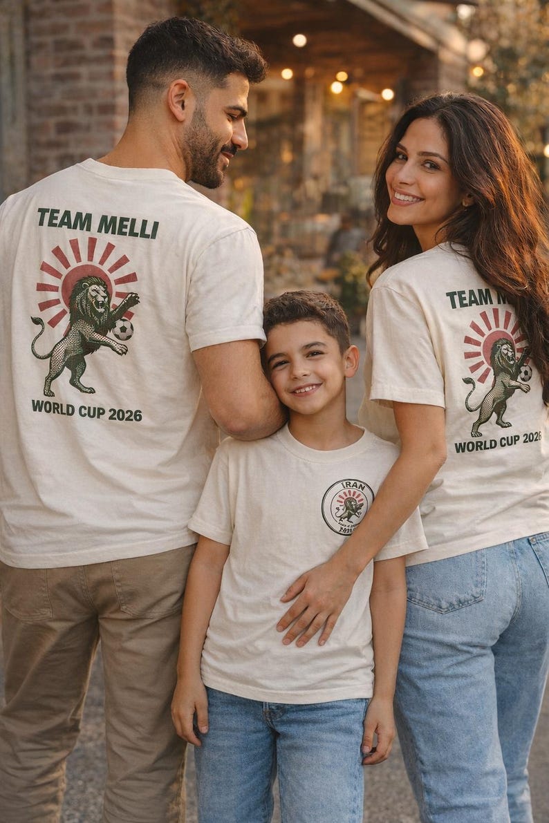 May include: Off-white short-sleeve t-shirts featuring a graphic on the back with a lion holding a soccer ball, a red sunburst, and the text TEAM MELLI WORLD CUP 2026. The smaller shirt features a circular logo on the chest with the text IRAN.