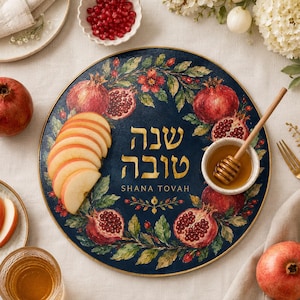 May include: A round dark blue serving plate with a gold rim, decorated with a pomegranate and leaf wreath. The center features the Hebrew text "שנה טובה" and the English text "SHANA TOVAH" in gold. A small bowl of honey with a wooden dipper sits on the plate.