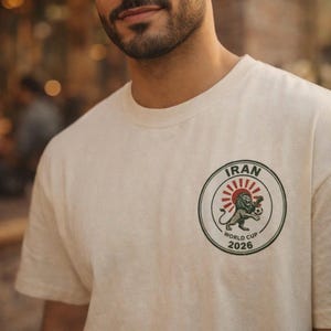 May include: A cream-colored t-shirt featuring a circular graphic on the chest. The design includes the text IRAN and WORLD CUP 2026, a red sunburst, and an illustration of a lion holding a soccer ball, all enclosed within a dark green border.
