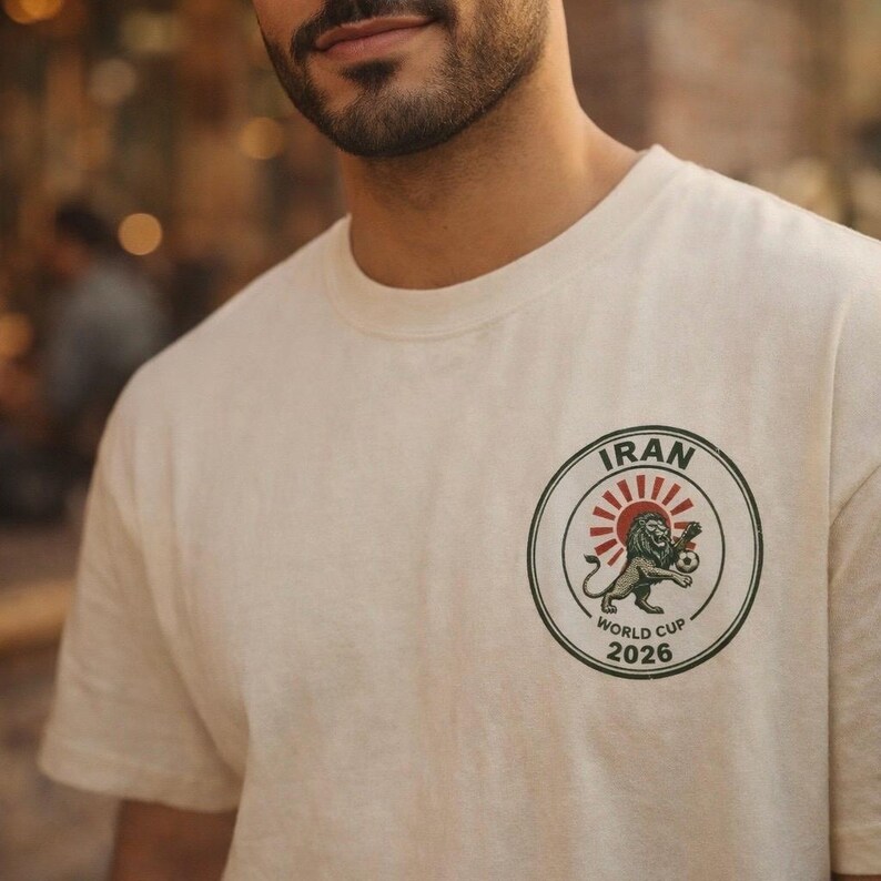 May include: A cream-colored t-shirt featuring a circular graphic on the chest. The design includes the text IRAN and WORLD CUP 2026, a red sunburst, and an illustration of a lion holding a soccer ball, all enclosed within a dark green border.