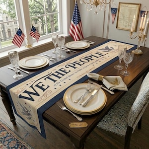 May include: A dining table set for a patriotic celebration. The table is adorned with a cream-colored runner featuring the text "WE THE PEOPLE" and a navy blue border. Place settings include white plates, silverware, and crystal glasses. American flags and decorative candles complete the scene.