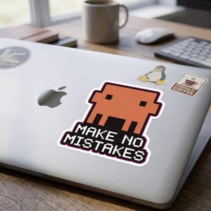 May include: A laptop with a sticker showing a pixelated orange creature and the words "MAKE NO MISTAKES". The sticker has a black and purple border. Other stickers and a coffee mug are also visible on the laptop.