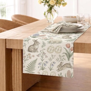 Puede incluir: Camino de mesa beige con temática de Pascua con un patrón repetido de conejos, huevos de Pascua y elementos florales. El camino de mesa está colocado sobre una mesa de madera, con platos, servilletas y un jarrón con flores.