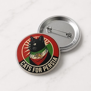 May include: A round button with a black cat wearing a scarf in the colors of the Iranian flag. The button has the text "CATS FOR PERSIA" in white. A second, unadorned button is visible in the background.