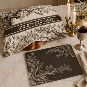 May include: A rectangular fabric challah cover featuring a black and white olive tree illustration and Hebrew text, paired with a matching dark rectangular placemat with white olive branch motifs and Hebrew text, alongside two brass candlesticks.