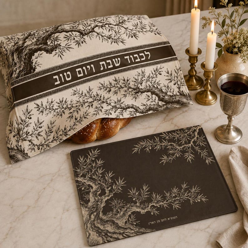 May include: A rectangular fabric challah cover featuring a black and white olive tree illustration and Hebrew text, paired with a matching dark rectangular placemat with white olive branch motifs and Hebrew text, alongside two brass candlesticks.