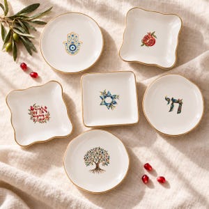 May include: A set of six white ceramic dishes with gold rims, featuring various designs including a hamsa, a pomegranate, a Star of David, Hebrew text, and a tree. The dishes are arranged with loose olives and pomegranate seeds.
