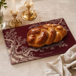 May include: A rectangular dark red serving board featuring a white illustration of an olive tree and Hebrew text. A braided challah bread topped with sesame seeds rests on the board. The board has a smooth finish and a decorative border design.
