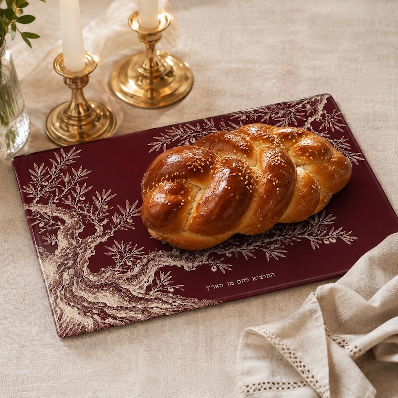 May include: A rectangular dark red serving board featuring a white illustration of an olive tree and Hebrew text. A braided challah bread topped with sesame seeds rests on the board. The board has a smooth finish and a decorative border design.