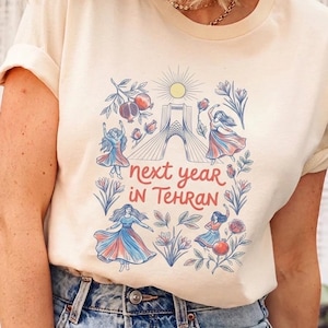 May include: Cream-colored t-shirt featuring a colorful graphic design. The design includes illustrations of dancing figures, pomegranates, flowers, and a bridge, with the text "next year in Tehran".