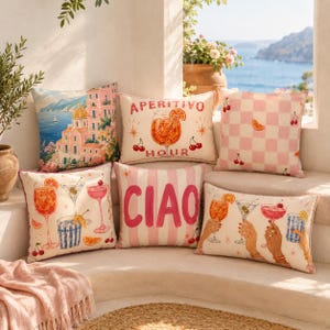 May include: A collection of five decorative throw pillows featuring coastal and cocktail-themed illustrations. Designs include a Mediterranean seaside town, a glass of orange spritz with the text APERITIVO HOUR, pink checkered patterns with cherries, and the word CIAO.