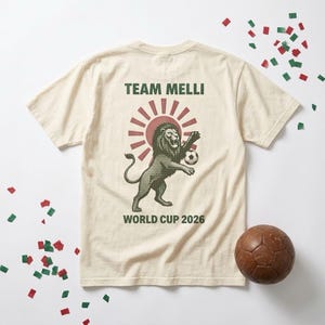 May include: A cream-colored short-sleeve t-shirt featuring a graphic on the back. The design includes a dark green lion holding a soccer ball, a red sunburst background, and the text "TEAM MELLI" and "WORLD CUP 2026" in dark green lettering.
