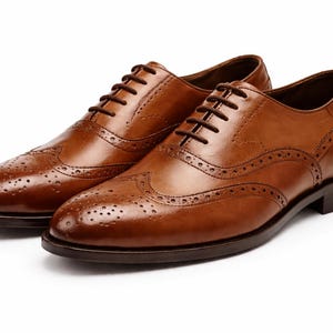 May include: A pair of brown leather Oxford shoes with brogue detailing. The shoes have a classic design with a lace-up closure and a dark brown sole. The leather has a polished finish, and the shoes are set against a white background.
