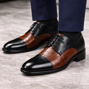 May include: Two-tone leather dress shoes. The shoes have a black toe and heel with a brown, textured centre. The shoes are laced up with black laces. The shoes are worn with black socks and navy blue trousers.