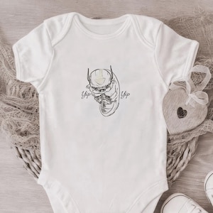 May include: White baby bodysuit featuring a black and white cartoon character illustration with the word "Yip" on either side. The bodysuit is shown with white baby shoes and a wooden heart.