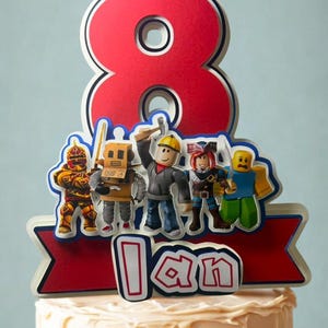 May include: A layered cake topper featuring a large red number eight, a group of five block-style character figures, and a red banner with the name Ian in white letters. The design uses red, blue, yellow, and grey colors with a multi-layered paper effect.