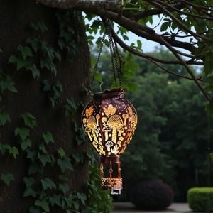 May include: A decorative, hanging lantern shaped like a hot air balloon. The dark brown metal lantern features a cut-out design of mushrooms and foliage, with a warm light emanating from within. It hangs from a chain attached to a tree branch.