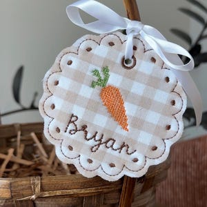 May include: A round, scalloped tag with a gingham pattern and the name "Bryan" embroidered in brown. An orange carrot with green leaves is embroidered on the tag. A white ribbon is tied to the top of the tag, which is attached to a woven basket.