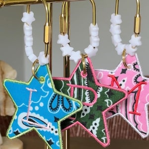 May include: Three star-shaped fabric keychains with embroidered letters I, D, and M. Each star features a different bandana print in blue, green, and pink. The keychains are attached to gold-toned metal rings decorated with white star-shaped beads.