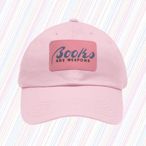 Puede incluir: Gorra de béisbol rosa claro con visera curva. Un parche rectangular en la parte delantera muestra el texto "Books are Weapons" en azul y gris. La gorra está hecha de tela suave y tiene una correa ajustable.
