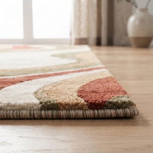 May include: A rectangular area rug featuring a tufted texture with a wavy, abstract pattern. The design consists of layered bands in shades of cream, muted olive green, tan, and terracotta orange, finished with a tightly bound edge in a neutral tone.