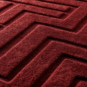 Op de afbeelding: Een dieprood tapijt met een verhoogd, gestructureerd geometrisch patroon. Het oppervlak bestaat uit een herhalend hoekig, doolhofachtig ontwerp dat wordt gecre&euml;erd door poolsecties die afsteken tegen de lagere, verzonken delen van het tapijt.