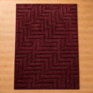 Op de afbeelding: Een rechthoekig vloerkleed in een diepe bordeauxrode kleur met een verhoogd, gestructureerd geometrisch patroon van in elkaar grijpende zigzaglijnen en rechte hoeken die een labyrintachtig ontwerp over het gehele oppervlak vormen.