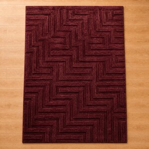 Op de afbeelding: Een rechthoekig vloerkleed in een diepe bordeauxrode kleur met een verhoogd, gestructureerd geometrisch patroon van in elkaar grijpende zigzaglijnen en rechte hoeken die een labyrintachtig ontwerp over het gehele oppervlak vormen.