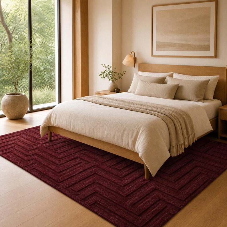Op de afbeelding: Een rechthoekig vloerkleed in een diepe bordeauxrode kleur met een verhoogd, geometrisch chevronpatroon. Het kleed ligt op een lichte houten vloer onder een houten bedframe met beige beddengoed, wat zorgt voor een contrast in textuur en kleur.
