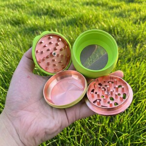 May include: A disassembled green and copper herb grinder. The grinder is held in a hand and is composed of several parts, including a grinding chamber, a screen, and a storage compartment. The background is a blurred green lawn.