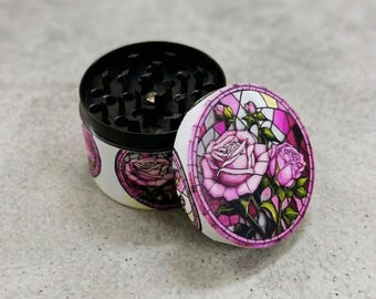 May include: A round, two-piece grinder with a black interior and a white exterior. The exterior features a stained-glass style design of pink roses and green leaves. The lid is open, revealing the grinding teeth.