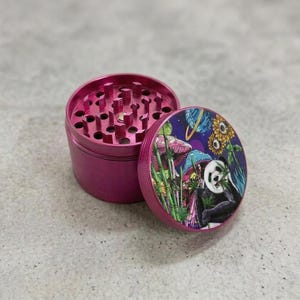 May include: A magenta-colored herb grinder with a lid featuring a colorful illustration of a panda, mushrooms, and sunflowers. The grinder is open, revealing the grinding teeth.