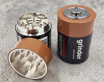 May include: A cylindrical herb grinder designed to resemble a D-cell battery with a black and brown exterior. The top cap is removed, revealing sharp metal teeth inside. The side features the text grinder, battery grinder, and warning labels.