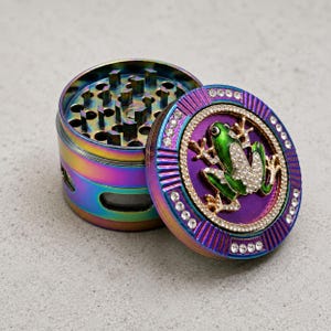 Puede incluir: Un grinder de hierbas multicolor e iridiscente, abierto. La tapa presenta un diseño de rana verde adornado con pedrería y un borde de pedrería púrpura. Los dientes del grinder son visibles.