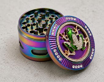 May include: A multi-colored, iridescent herb grinder with a lid featuring a green frog design adorned with rhinestones. The grinder is open, revealing the grinding teeth. The lid is purple with a rhinestone border.