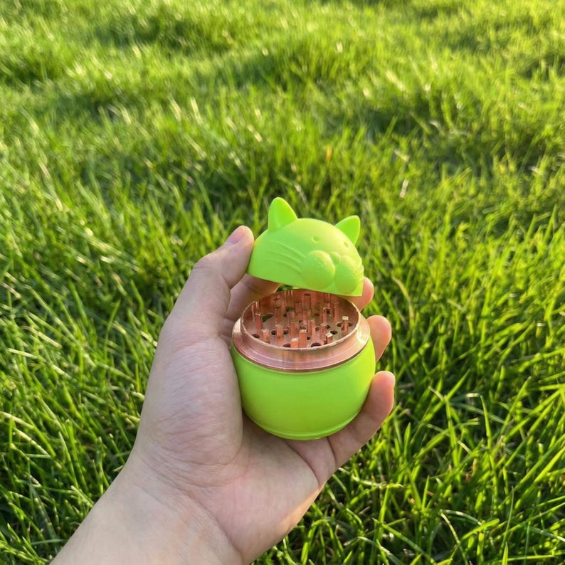 May include: A lime green cat-shaped herb grinder with a copper-colored grinding chamber. The top of the grinder is open, revealing the grinding teeth. The grinder is held in a hand against a backdrop of green grass.