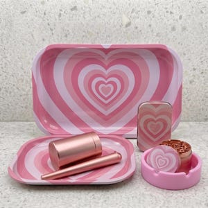 May include: A rectangular rolling tray, a smaller square tray, a metal tin, a cylindrical container, a slender tube, and a circular grinder, all featuring a pink and white concentric heart pattern, alongside a solid pink circular ashtray.