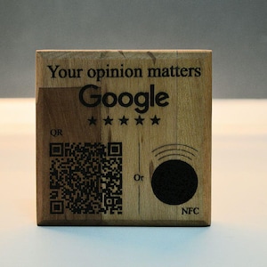 Può includere: Un blocco di legno con il logo Google, cinque stelle e la scritta "Your opinion matters". È dotato di un codice QR e di un simbolo NFC. Il blocco è in legno marrone chiaro.