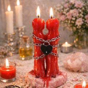May include: A red candle shaped like two figures embracing, bound by a chain and a heart-shaped lock. The candle sits on a pinkish base, with a lit flame at the top of each figure. Additional candles and decorative items surround it.