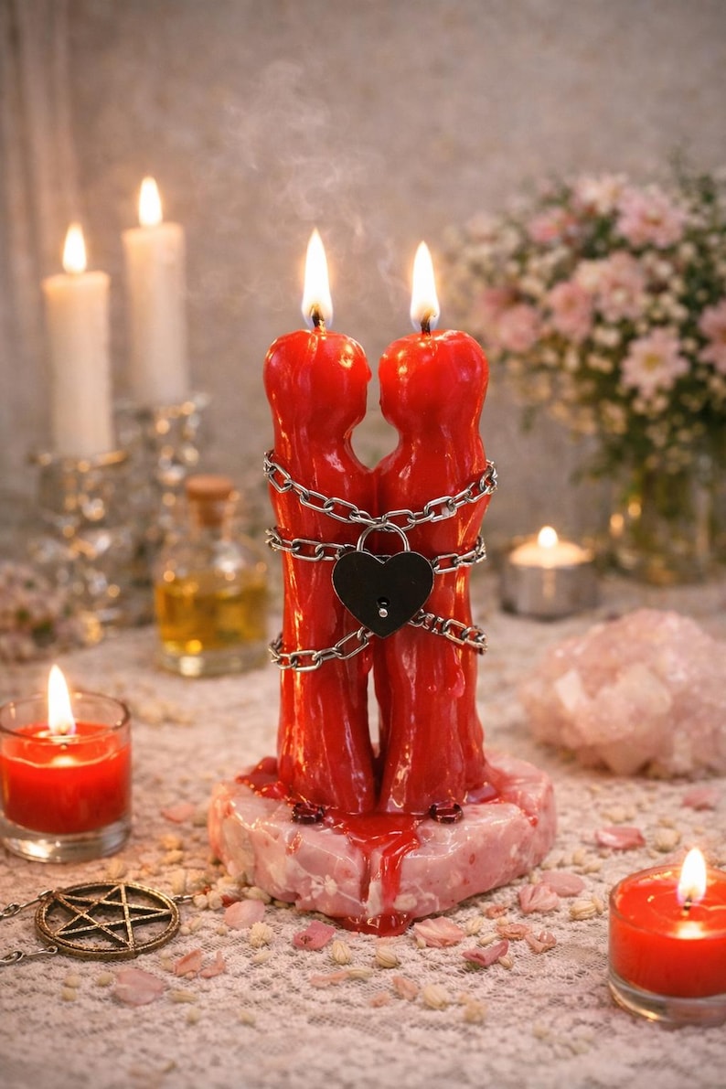 May include: A red candle shaped like two figures embracing, bound by a chain and a heart-shaped lock. The candle sits on a pinkish base, with a lit flame at the top of each figure. Additional candles and decorative items surround it.