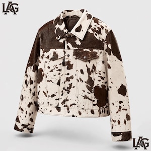 May include: A long-sleeve jacket featuring a brown and white cow print pattern throughout. The jacket includes a standard collar, a front button closure, and two chest pockets with buttoned flaps. The letters LAG are visible in the corners.
