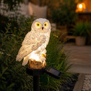 Peut inclure: Lampe solaire de jardin en forme de hibou blanc, avec des yeux jaunes et une surface texturée. Le hibou est posé sur une base décorative, montée sur un piquet noir avec un petit panneau solaire. La lumière est allumée, créant une lueur chaleureuse.