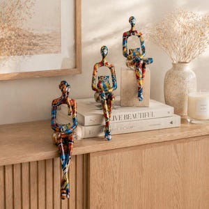 Peut inclure: Trois sculptures abstraites de figures assises aux motifs colorés. Les sculptures sont placées sur un meuble en bois, avec des livres, un vase de fleurs séchées et une bougie à proximité. Les figures ont un design moderne et artistique.