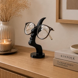 Peut inclure: Un porte-lunettes décoratif noir en forme de visage, tenant une paire de lunettes. Le support est posé sur une surface en bois, avec un vase, une bougie et des livres en arrière-plan. Les lunettes ont une monture dorée.